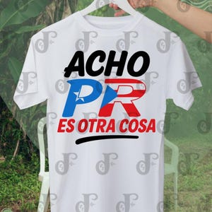 May include: White t-shirt with the text "ACHO PR ES OTRA COSA" in black, blue, and red. The design incorporates the Puerto Rican flag. The shirt is hanging on a white hanger against a blurred background.