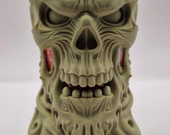 May include: An olive-green, 3D-printed skull-shaped drinks can cooler. The cooler features detailed facial features, including a nose, eye sockets, and teeth. A red can is placed inside the cooler.