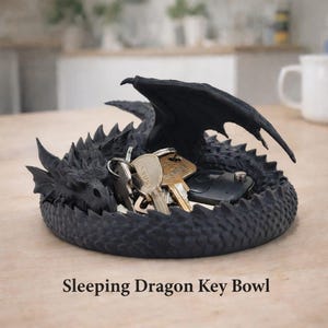 Op de afbeelding: Een zwarte draakvormige sleutelschaal met een slapend drakenontwerp. De schaal bevat sleutels en een autosleutelhanger. De tekst "Sleeping Dragon Key Bowl" staat onderaan. De draak heeft gedetailleerde schubben en vleugels.