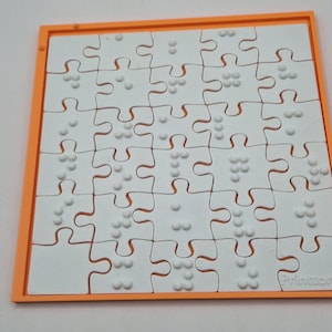 Puede incluir: Un rompecabezas blanco con bordes naranjas y puntos en braille en cada pieza. El rompecabezas es cuadrado y tiene un patrón de cuadrícula. La palabra "Printor" es visible en la esquina inferior derecha del rompecabezas.