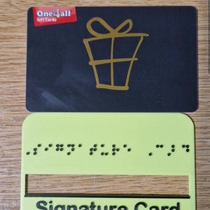 May include: A black One4all gift card featuring a gold outline of a gift box with a bow, positioned above a light green plastic signature card guide. The green card features embossed Braille dots and the black text Signature Card.
