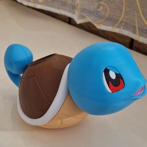 May include: A blue, brown, and tan Squirtle figurine. The figurine has a blue head and tail, a brown shell, and a tan body. The eyes are black with red accents, and the mouth is pink. The figurine is a 3D-printed item.