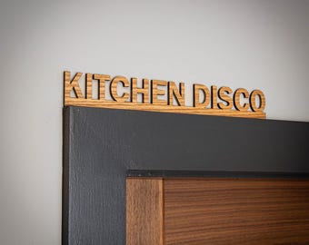 May include: A wooden sign with the text KITCHEN DISCO in a sans-serif font, featuring a natural light brown wood grain finish. The sign is positioned horizontally along the top edge of a dark gray door frame with a brown wood panel door.