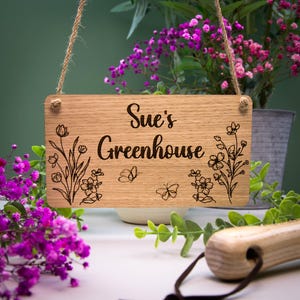 K&ouml;nnte beinhalten: Ein rechteckiges, h&auml;ngendes Schild aus hellbraunem Holz mit nat&uuml;rlicher Maserung. Der Text "Sue's Greenhouse" ist mittig in einer dunklen Schreibschrift eingraviert. Das Schild zeigt eingravierte Illustrationen von Wildblumen und zwei Schmetterlingen.