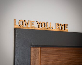 May include: A light brown wooden sign with the text LOVE YOU, BYE cut out in a sans-serif font. The sign is positioned horizontally along the top edge of a dark gray and wood-toned door frame, featuring a visible wood grain texture throughout.
