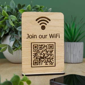 May include: A rectangular light-coloured wooden sign with rounded corners, featuring a dark brown laser-engraved Wi-Fi symbol, the text "Join our WiFi," and a square QR code. The sign is mounted on a thin, dark brown rectangular base.