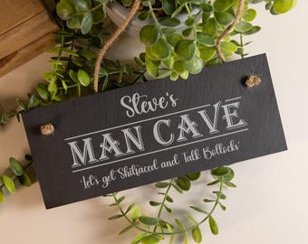 May include: A rectangular black slate sign featuring the engraved white text "Steve's MAN CAVE" and "Let's get Shitfaced and Talk Bollocks". The sign includes two holes with a natural jute twine rope for hanging, displaying a textured surface.
