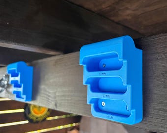 May include: Two bright blue, tiered training holds for climbing, mounted to a dark wooden beam. Each hold features three distinct horizontal slots labeled 30 mm, 20 mm, and 10 mm, with visible silver screws securing them to the wood surface.