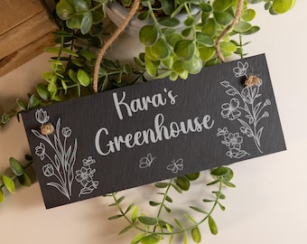 May include: A rectangular black slate sign hanging by a jute rope, featuring the white engraved text "Kara's Greenhouse" in the center. The sign is decorated with white line art illustrations of flowers and butterflies on both sides.