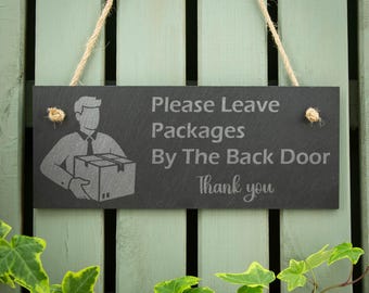 May include: A rectangular dark gray slate sign hanging from a jute rope. The sign features an engraved graphic of a person holding a package and the text: Please Leave Packages By The Back Door Thank you. The surface has a natural stone texture.