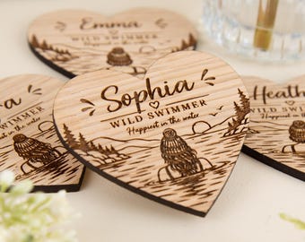 May include: Heart-shaped wooden coasters featuring laser-engraved designs. Each piece displays a name, the text WILD SWIMMER, and the phrase Happiest in the water, accompanied by an illustration of a person in a beanie swimming in a lake.