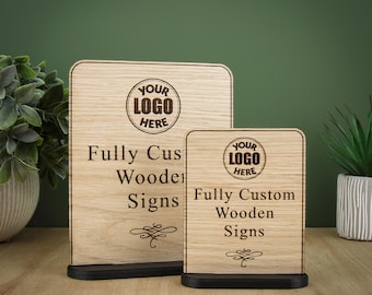 Könnte beinhalten: Zwei rechteckige Schilder aus hellem Holz mit abgerundeten oberen Ecken, die jeweils auf einem schwarzen Sockel stehen. Die Schilder tragen den eingravierten Text "YOUR LOGO HERE", "Fully Custom Wooden Signs" und ein Zierelement unten.