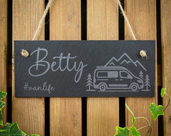 May include: A rectangular black slate hanging sign with a jute rope. The sign features the engraved name Betty in a script font, the hashtag #vanlife, and a graphic illustration of a camper van surrounded by pine trees and mountain outlines.