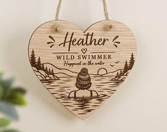 May include: A heart-shaped wooden plaque with a jute rope hanger. The surface features a laser-engraved illustration of a person in a beanie swimming in a lake, with pine trees and a sun. Text reads: Heather, WILD SWIMMER, Happiest in the water.