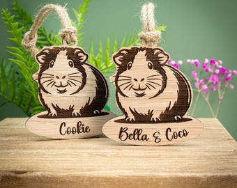 May include: Two wooden guinea pig ornaments with a natural wood grain finish. Each ornament features a detailed engraving of a guinea pig and a name: "Cookie" and "Bella & Coco". They have twine loops for hanging.