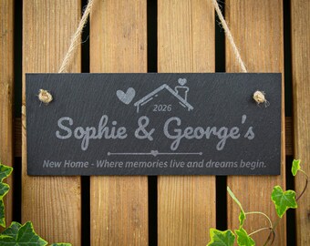May include: A rectangular black slate sign hanging by a jute rope. The sign features an engraved heart, a house outline, the year 2026, the names Sophie & George's, and the text New Home - Where memories live and dreams begin.