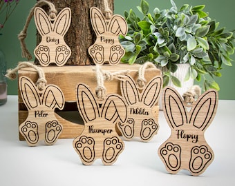 May include: Wooden Easter bunny ornaments with names like "Daisy" and "Poppy" are displayed. Each ornament features a bunny shape with paw prints and a jute string for hanging. The ornaments are made of light-colored wood.
