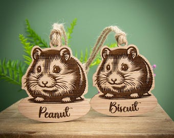 Könnte beinhalten: Zwei Holzornamente in Hamsterform mit eingravierten Details. Jedes Ornament zeigt ein Hamsterdesign und einen Namen: "Peanut" und "Biscuit". Sie hängen an Schnurschlaufen.