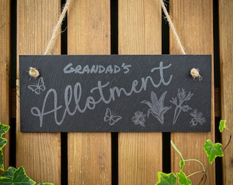 May include: A rectangular black slate sign hanging by a jute rope. The sign features the engraved text "GRANDAD'S Allotment" in a script font, accompanied by etched illustrations of two butterflies, a corn cob, and various garden vegetables.