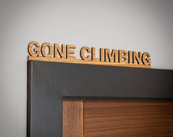 May include: A wooden sign with a natural grain finish featuring the text GONE CLIMBING in a bold, sans-serif font. The sign is positioned horizontally along the top edge of a dark-painted door frame, showcasing a clear wood texture.