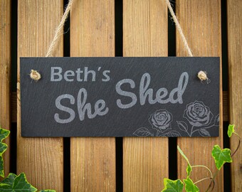 May include: A rectangular black slate sign featuring the engraved text Beth's She Shed in a light gray font. The sign includes an etched rose design on the right side and is suspended by a natural jute twine rope threaded through two holes.