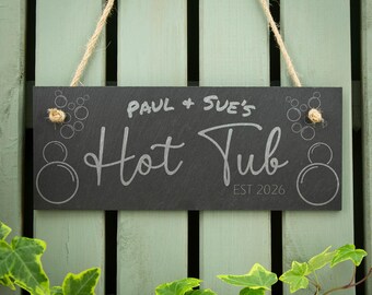 May include: A rectangular black slate sign hanging from a jute rope. The sign features the engraved text "PAUL + SUE'S Hot Tub" and "EST 2026" in a light gray color. Decorative etched circles resembling bubbles are positioned on the left and right sides.