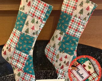 May include: Two patchwork Christmas stockings featuring a repeating pattern of red and green plaid, beige fabric with small green and red Christmas trees, and teal fabric with gold reindeer silhouettes. A small white rectangular tag reads Theodore.