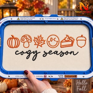 Könnte beinhalten: Ein blauer 20,32x33,02 cm Stickrahmen mit cremefarbenem Stoff, darauf orange gestickte Symbole von Kürbis, Pilz, Blatt, Smiley, Tortenstück und Eichel. Darunter ist der Text cozy season in schwarzer Schreibschrift aufgestickt.