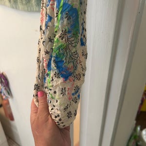 Può includere: Sciarpa color crema con motivo floreale e a stella, impreziosita da spruzzi di vernice blu, verde e rossa. È visibile il tessuto leggero e strutturato della sciarpa. La sciarpa è tenuta in una mano.