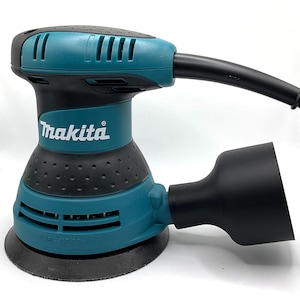 Pode incluir: Lixadeira orbital aleatória Makita, teal e preta. A lixadeira tem uma alça preta e uma porta de coleta de poeira preta. O logotipo da Makita é visível em branco. Base redonda e cabo de alimentação preto.