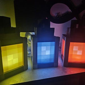 May include: Three Minecraft-themed lanterns, each with a different colored light: yellow, blue, and orange. The lanterns are made of a black material and have a square shape.