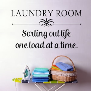 May include: A laundry room scene with the text "LAUNDRY ROOM" and "Sorting out life one load at a time." An ironing board holds a white iron and a stack of folded towels. A wicker basket filled with folded towels sits nearby.