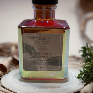 May include: A Smokey-White Cedarwoods reed diffuser in a square iridescent glass bottle with a black cap. The label reads "Back Settlement Aroma, St David ME, EST 2024". The bottle contains 4 fl oz of fragrance oil and sits on a wooden surface.