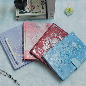 May include: Four different journals with floral designs and the text "love letter from a" on the front covers. The journals are in shades of blue, purple, pink, and red.