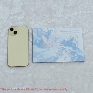 May include: A white iPhone 15 is next to a blue and white card with a pink butterfly and the text "The flapping wings of butterflies are resonating with freedom".