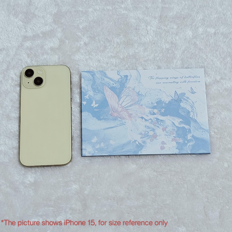 May include: A white iPhone 15 is next to a blue and white card with a pink butterfly and the text "The flapping wings of butterflies are resonating with freedom".