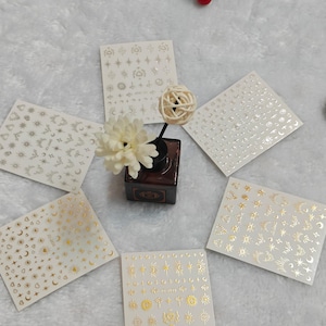 Pode incluir: Adesivos de nail art com desenhos celestiais. Os adesivos são dispostos em torno de um pequeno objeto decorativo e uma flor. Os adesivos têm acabamentos metálicos dourados e prateados, com sóis, luas, estrelas e outras formas celestiais.