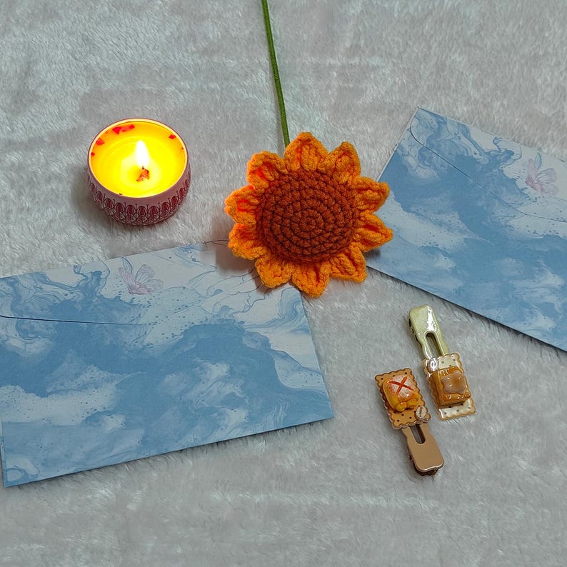 May include: A blue and white marbled envelope, a crocheted orange sunflower, a small candle in a pink holder, and two gold hair clips with a brown and white pastry design.