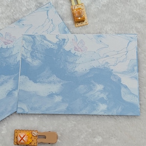 May include: Two blue envelopes with a marbled pattern and a white butterfly design. The envelopes are folded and ready to be used for sending letters or cards.