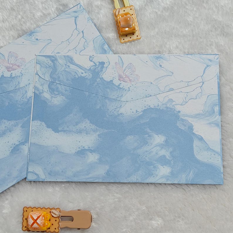May include: Two blue envelopes with a marbled pattern and a white butterfly design. The envelopes are folded and ready to be used for sending letters or cards.