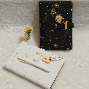 May include: Two decorative journals with celestial designs. One is black with gold accents, a heart-shaped lock, and a chain. The other is white with gold accents and a pen. Both have star and constellation patterns.