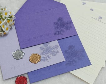 Może przedstawiać: Zestaw artykułów papierniczych z fioletowymi i lawendowymi kopertami i notatnikami z kwiatowym wzorem róży. Zestaw zawiera papier w linie, pieczęcie woskowe w kolorze srebrnym, złotym i czerwonym oraz tekst "T. MENISHPBJSP".