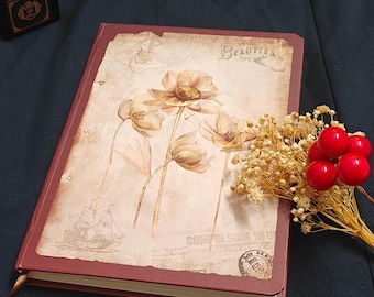 May include: A maroon journal with a floral design on the cover. The cover features a vintage-style illustration of flowers and a ship. A small bouquet of dried flowers and red berries rests beside the journal.