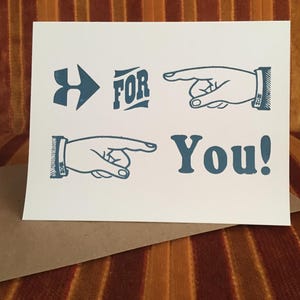 May include: A white greeting card with blue text and illustrations. The card reads "H FOR You!" with a hand pointing towards the words. A brown envelope is beneath the card.