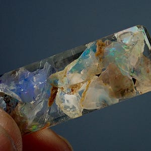 May include: A clear glass tube filled with iridescent opal chips. The opal chips are various shades of blue, green, and white. The tube is approximately 2 inches long.