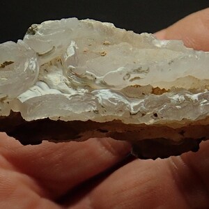 May include: A close-up of a layered stone, displaying a mix of white, beige, and light brown colours. The stone's surface has a wavy, textured appearance, with visible crystalline structures. The overall aesthetic suggests a natural, geological specimen.