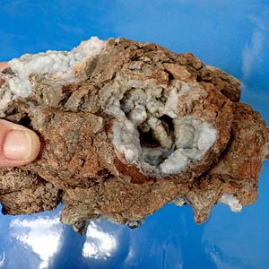 May include: A rough, brown rock with a cavity filled with white and light blue crystals. The crystals are clustered together and have a sparkly appearance.