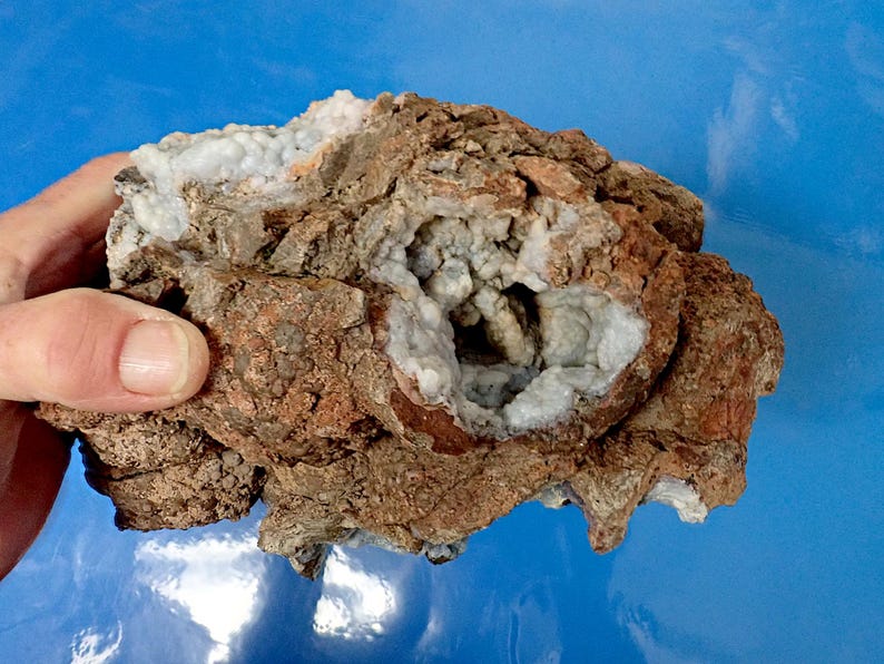 May include: A rough, brown rock with a cavity filled with white and light blue crystals. The crystals are clustered together and have a sparkly appearance.