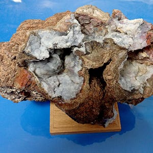 May include: A large, irregularly shaped rock with a hollow centre filled with white crystals. The rock is brown and grey with reddish-brown accents. It is displayed on a wooden base.