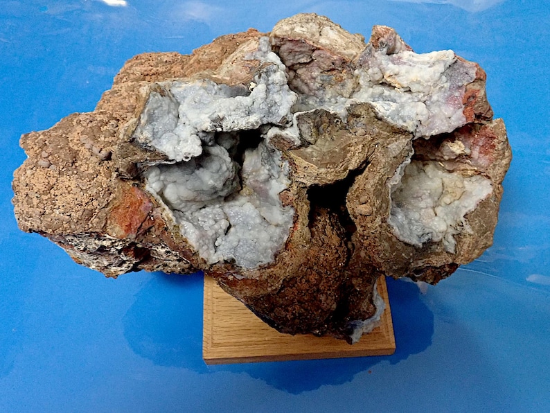 May include: A large, irregularly shaped rock with a hollow centre filled with white crystals. The rock is brown and grey with reddish-brown accents. It is displayed on a wooden base.